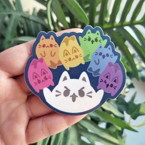 a photo of a glitter sticker design held in front of a leafy green plant. The sticker design shows several cute stylized ghost cats in a rainbow of different colors and poses.