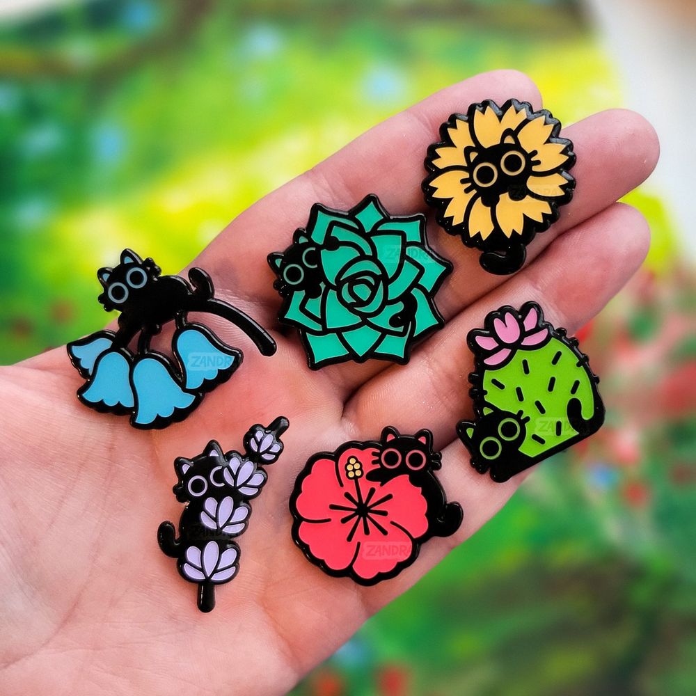 A set of black dyed enamel pins each featuring a cat peeking out of a different flower or plant. Bluebells, Succulents, Sunflowers, lilacs, hibiscus, and cactus plants are featured. 