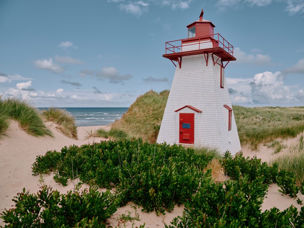 St. Peter’s Lighthouse, Prince Edward Island.

A small white wooden heritage lighthouse with red trim stands nestled among sand dunes and beach grasses on the north shore of Prince Edward Island at St Peter's. The sky is clear and blue, with the Gulf of St. Lawrence visible just beyond the dunes. The scene is bright, breezy, and coastal.
