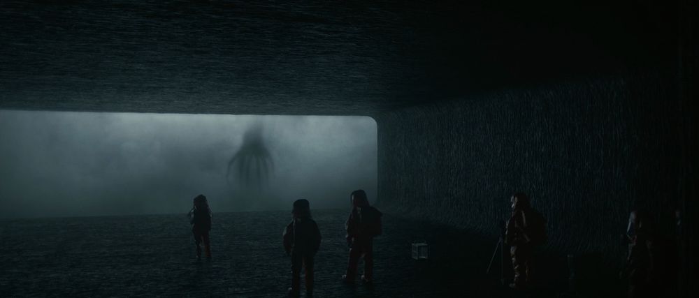 A still from the film "Arrival". A group of humans in protective suits stand in a dark rounded-rectangular tunnel that ends in a glass wall. Beyond the wall, a large octopus-like shadow looms in thick mist. 