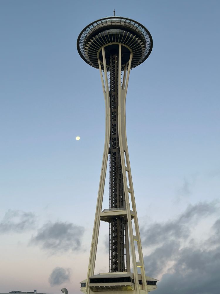 Early morning sky, the space needle, and a waning nearly-full moon to its left. Wisps of clouds on the left consolidate to darker, thicker clouds on the right (to the north).