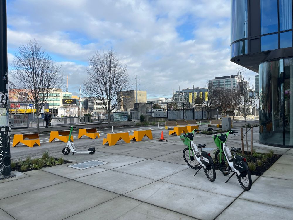 A city block with no car access, aside from alleyway entry halfway down. Yellow benches take up half the street while a bicycle lane occupies the other. A couple LimeBikes and a scooter are appropriately tucked along the sides of a wide sidewalk. A skate park is visible behind the street. 

There is no longer a crosswalk sign here because one is no longer needed.