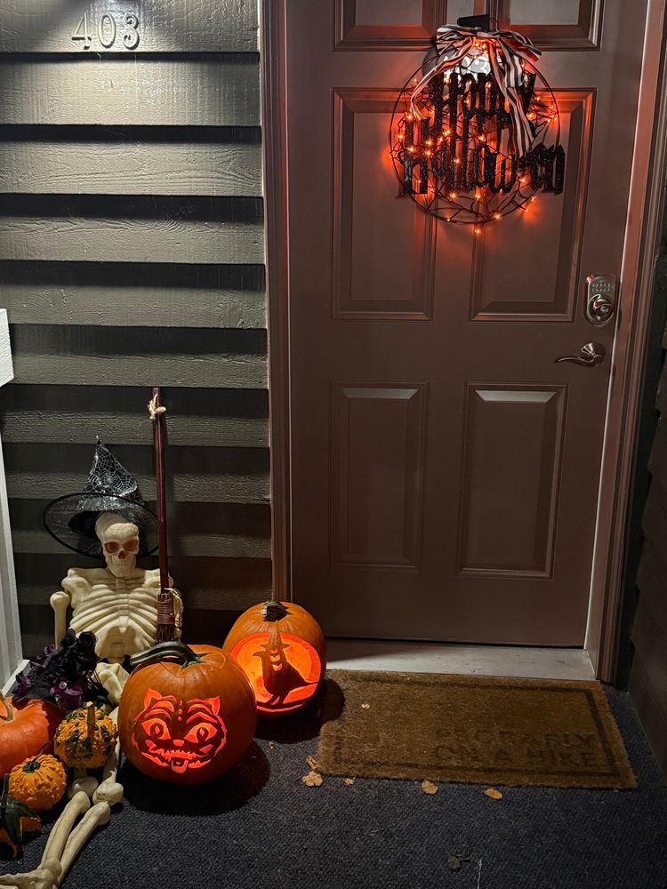 Picture of a front door to an apartment. To the left, there’s a plastic skeleton wearing a witch’s hat and holding a broomstick surrounded by pumpkins 2 of which are carved jack-o-lanterns of Derpy and Sussy from “KPop Demon Hunters”. The door has a black spider web “Happy Halloween” sign lit up by orange lights. 