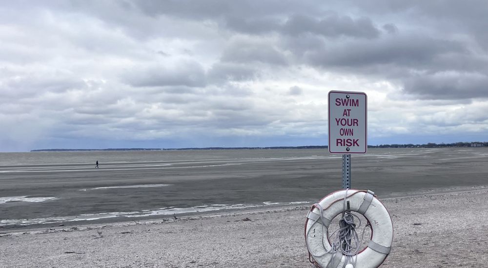 Very low water along a lakeshore, with a person walking where there would normally be at least 4-5 more feet of water. A life preserver in the foreground is attached to a sign that says "SWIM AT YOUR OWN RISK".