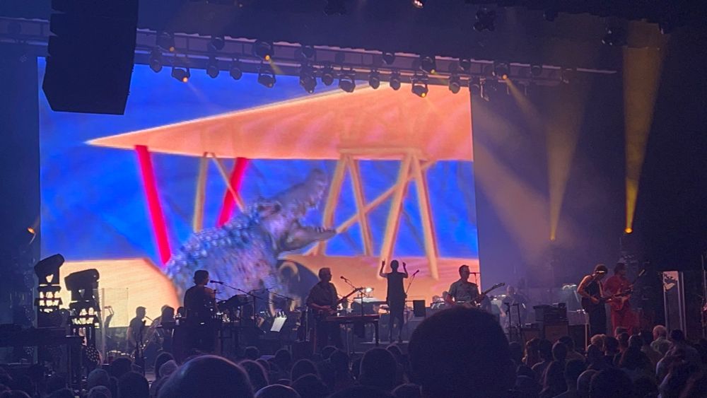King Gizzard & the Lizard Wizard playing on stage with Sarah Hicks conducting the Chicago Philharmonic. Background screen shows an alligator flying a yellow plane on a blue background.
