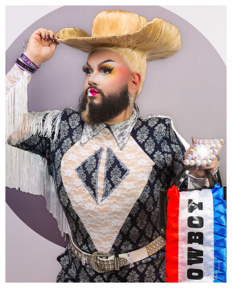 A photo of a drag queen wearing an elaborate blue and white outfit with fringe and a wig in the shape of a cowboy hat. 