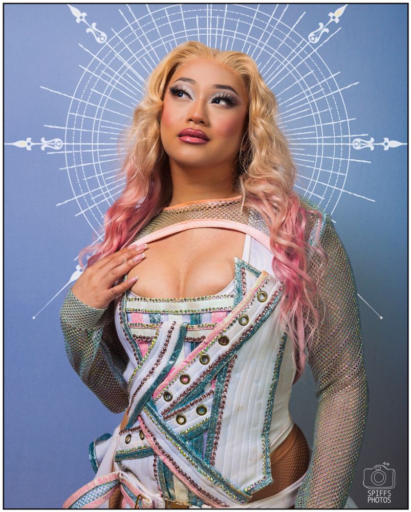 A picture of a drag queen in a white, pink and blue corset looking up. 