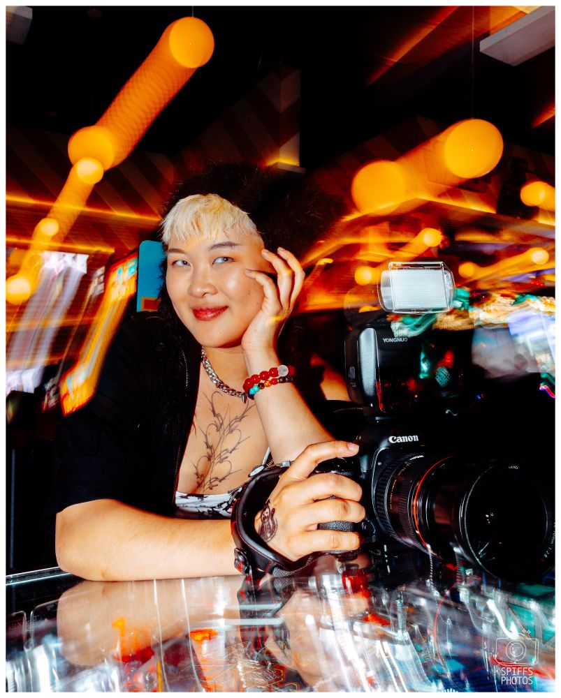 A picture of an Artist leaning on a pinball machine holding a camera and smiling. 