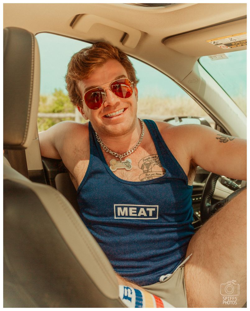 My friend wearing a blue tank top, short grey shorts, glasses sitting in a car, looking at the camera and smiling. 