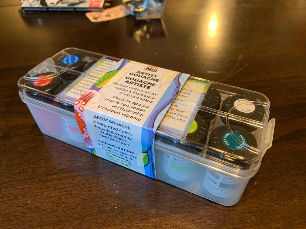 on a wooden table surface sits a clear plastic box full of small jars of paint. The paper label wrapped around the box reads:

Niji

Artist Gouache
Gouache Artiste
Opaque watercolor for design and illustration
12 vibrant colors

Gouache opaque
pour la conception
et l’illustration
12 couplers vibrant

Then on the front side of the box:
ARTSI GOUACHE
12 Saturated Colors
Smooth and Creamy
Matte Finish 
Fade Resistant

GOUACHE ARTISTE
12 Couleurs Saturées
Lisse and Crémeux
Finition Mate
Resistant a la
Décoloration

more art supplies are also visible in the far background.