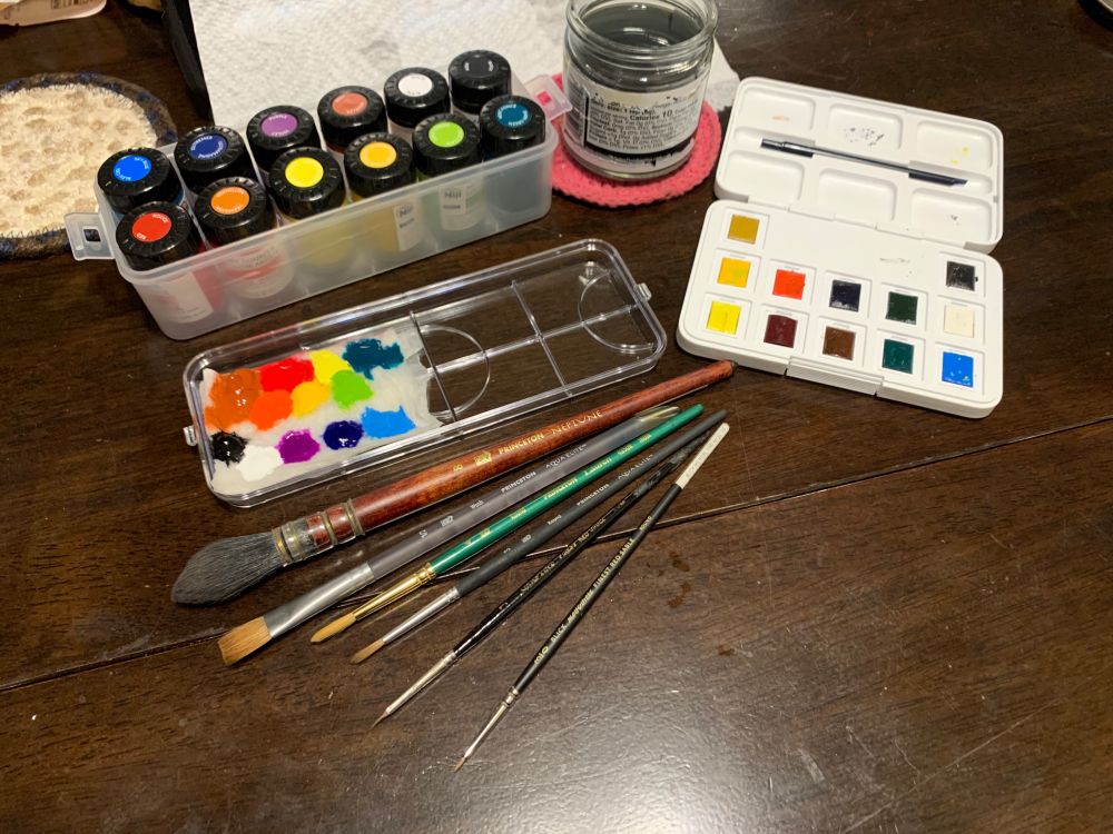 Various art supplies sit on a wooden table: A plastic box filled with 12 tiny jars of different colors of gouache paint, a plastic palette with a dab of each of the colors laid out on a dampened paper towel, another dry paper towel, a small jar of paint water, a Van Gough portable set of 12 watercolor paint cakes with an attached palette and six watercolor brushes in a variety of sizes and shapes, most of them round with one flat brush. 