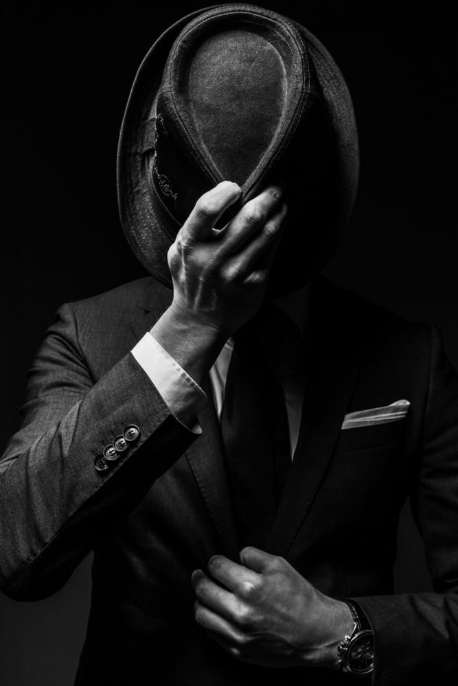 A dark, black and white photograph of a man wearing a classy suit and holding a hat in front of his face. The shot has a high contrast, making the light parts stand out and the dark extra black. The shot gives you a 60s vibe with the style of the hat and the classy way the man is dressed.