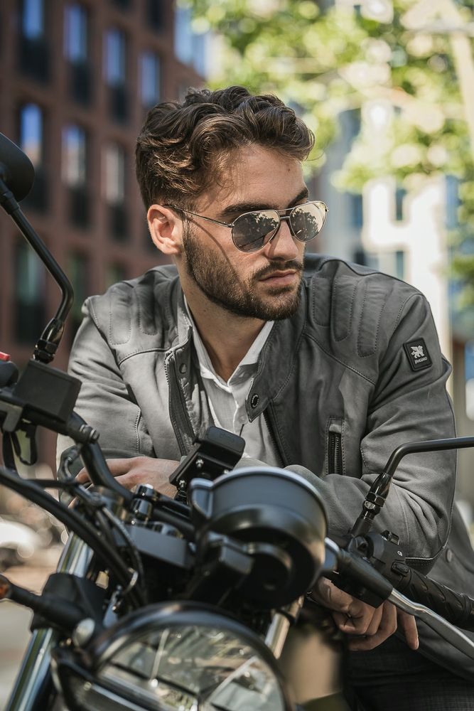 A though guy sitting on a motorcycle of the brand Moto Guzzi. He is wearing a grey leather jacket and dark sunglasses. He has a beard and brown wavy hair. He looks of to the right as he leaning over the stearing wheel of his motorcycle. This look is inspired by a Vanguard Clothing commercial. A cool Dutch brand of men's clothing.