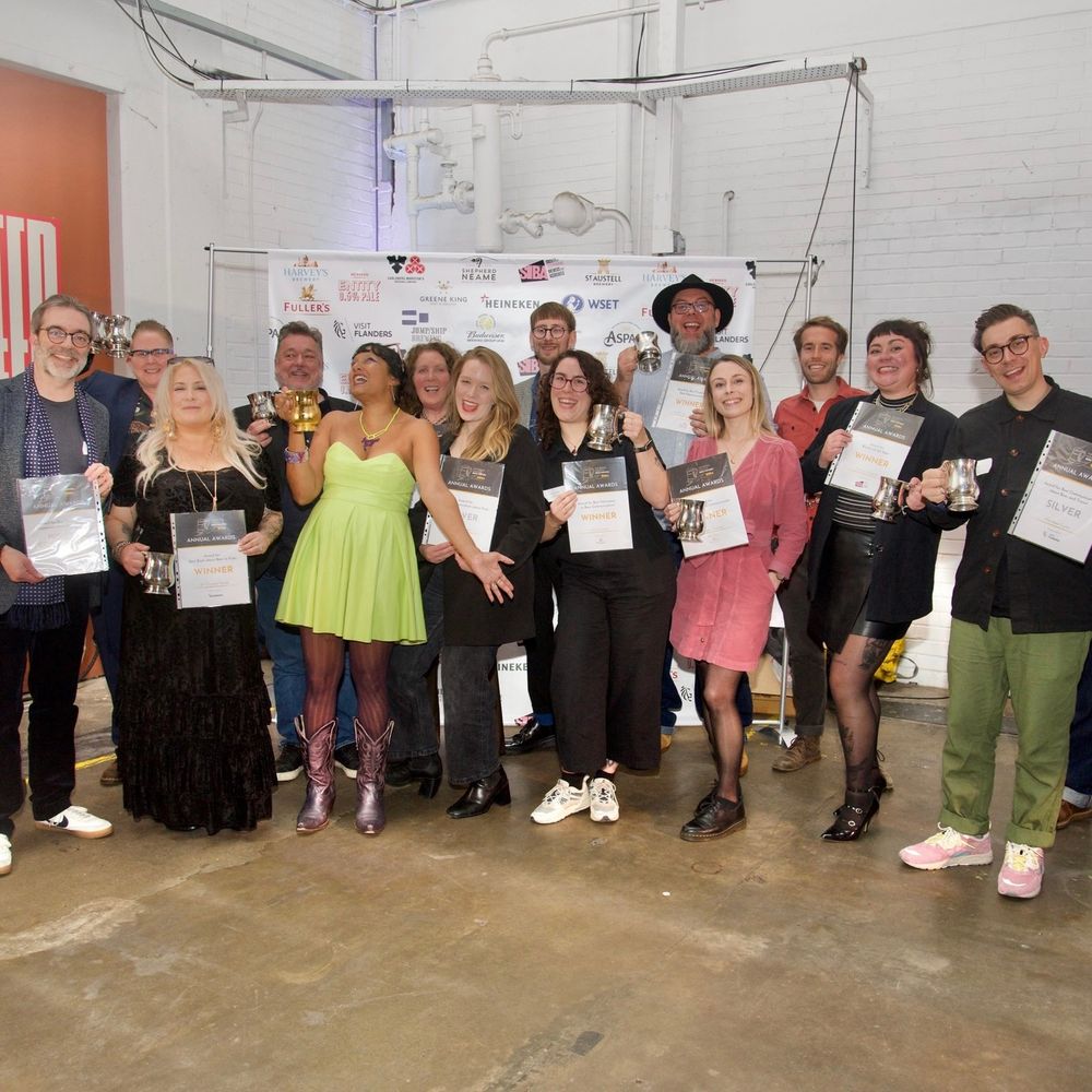 A group of Guild members pose together indoors in front of a branded backdrop at the British Guild of Beer Writers Awards 2024. They are smiling and holding silver tankards and framed certificates. The setting is Big Penny Social, an industrial-style venue with white brick walls and visible pipes.