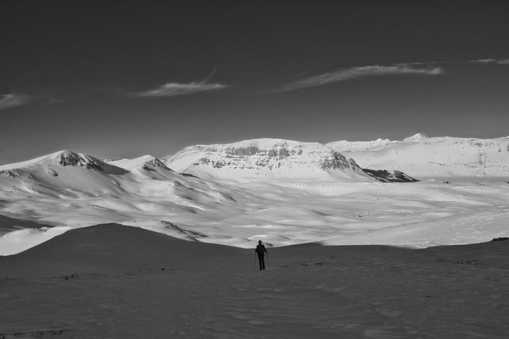 Un escursionista attraversa un altopiano innevato. Sullo sfondo, montagne si stagliano contro un cielo scuro con nuvole sottili.