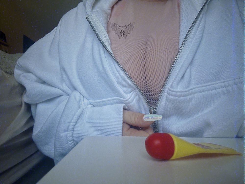 soul (white, afab creator) is photographed in a white, zip up hoodie that is zipped half-way down, to expose a sheer top and cleavage. her hand is holding the zipper, about to pull it down more.