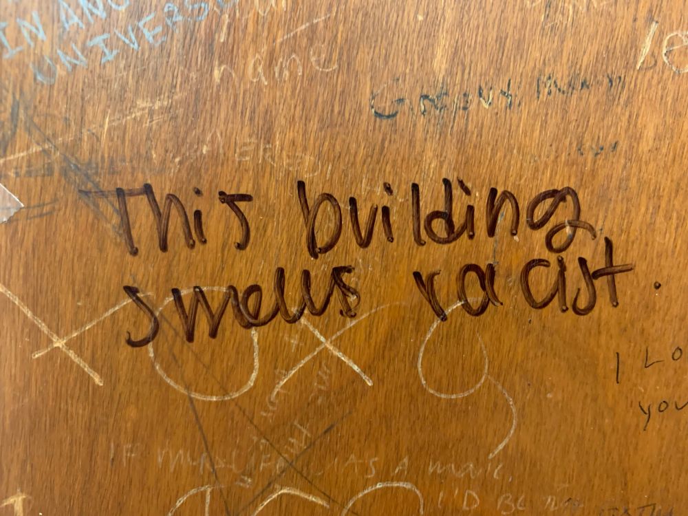 A wooden door with the phrase this building smells racist written on it in black sharpie. 