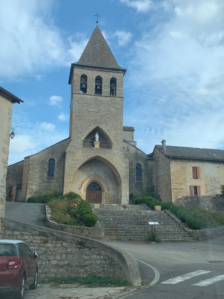 A stone church at the top of a set of stairs. It has a recessed porch leading to the front door, and the front of the church rises up flat into a wide bell tower. The bell tower has three arched openings where bells are visible. Above the porch is a second smaller recessed niche with a statue inside it.