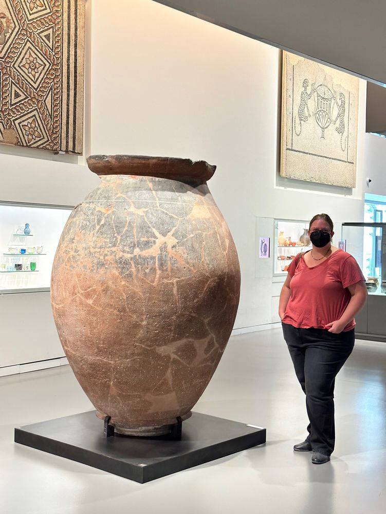 Photo of a huge terra cotta dolium or storage jar, which is mounted on a black platform. A woman wearing black jeans, a red t shirt, glasses and a black mask is standing next to it, and it is at least a foot taller than her. There are mosaics mounted on the wall behind them.