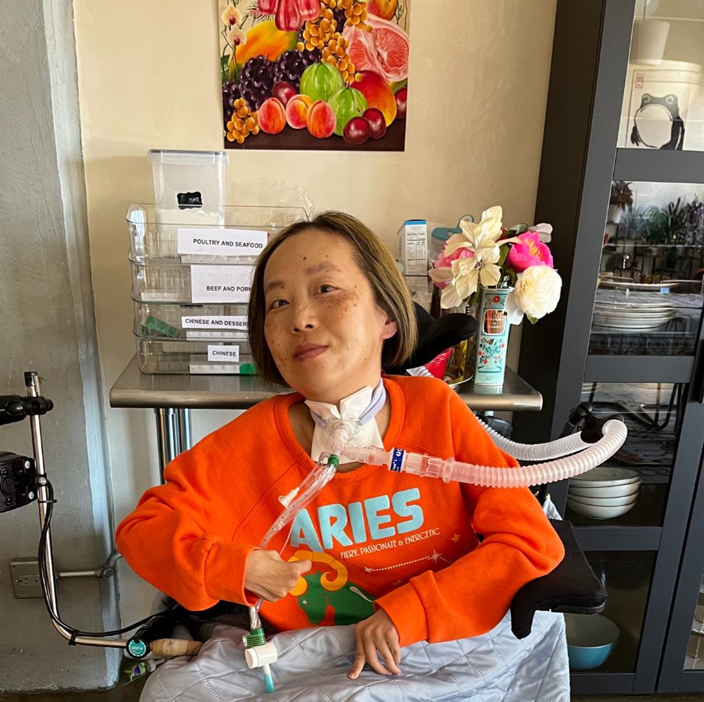 Asian American woman with blonde hair in a wheelchair. She has a tracheotomy at her neck with a tube attached to a ventilator She is wearing an orange sweatshirt that says ARIES. There is a pale blue blanket over the lap