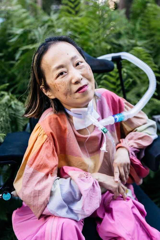 Asian American woman in a wheelchair with a tracheostomy at her neck connected to a ventilator. She’s wearing a pink plaid shirt, pink pants, and a magenta lip color. She is smiling and behind her are a bunch of tall prehistoric looking plants. Photo credit: Allison Busch Photography.