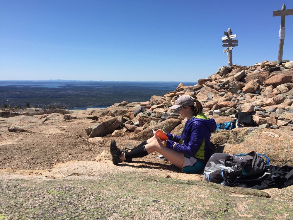 a scientist sits on the summit of a mountain in Acadia National Park, Maine. She is wearing a purple jacket and blue baseball cap, writing in an orange notebook. Behind her, the view stretches out across Mount Desert Island forests into the Gulf of Maine and distant islands.