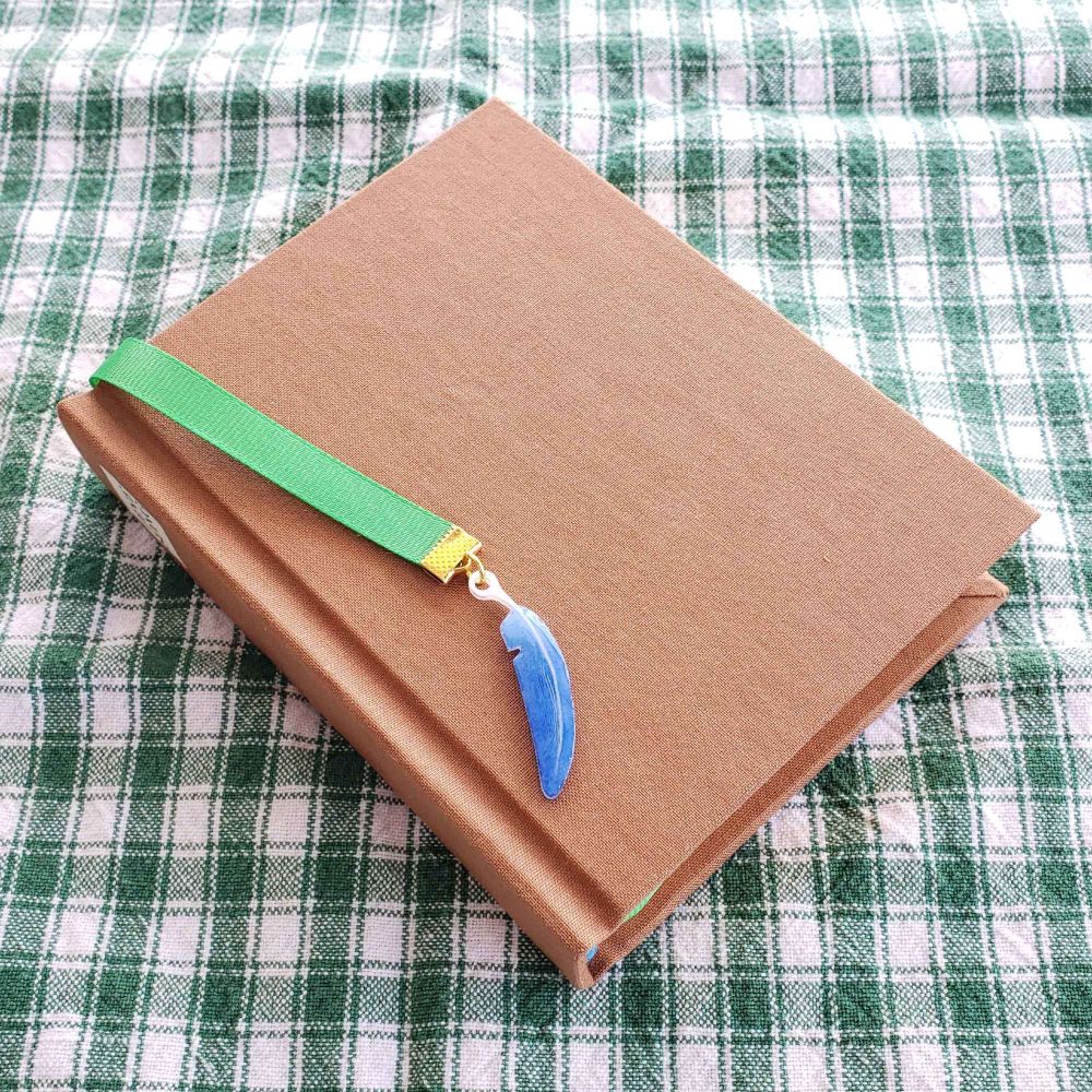 Brown book laying on green and white cloth. Attached to it is a green ribbon bookmark with a blue feather charm on the end.
