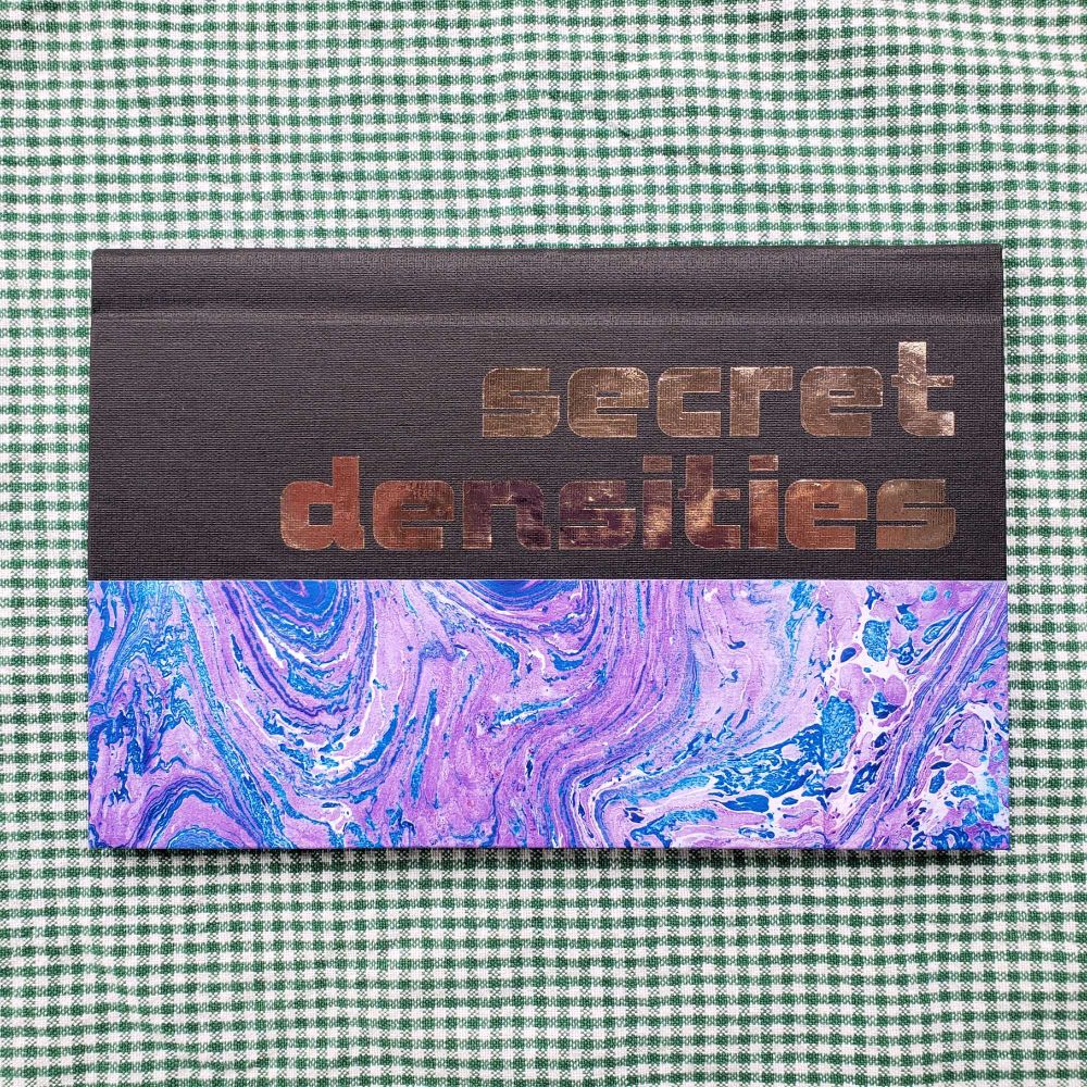 Hardcover pamphlet bind. The spine and part of the cover is covered in black bookcloth. Purple and blue marbled paper cover the rest. The title, "secret densities" is there in metallic iron-on.
