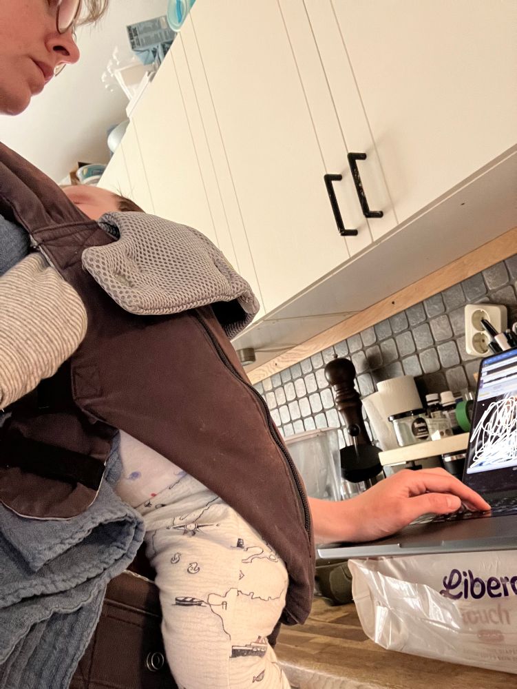 Person standing at counter with baby harness on, also typing on computer which is elevated using a pack of diapers.