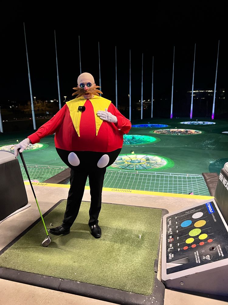 Dr. Eggman standing holding a golf club at a TopGolf driving range. It’s nighttime in the photo.