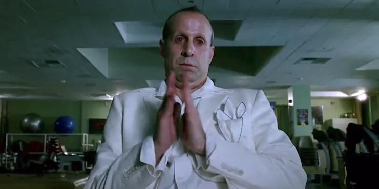 Peter Stormaire as Lucifer, a balding white middle-aged man in a white suit sitting in an office gym. He’s clapping his hands sarcastically and has a look like he’s patronizing you.