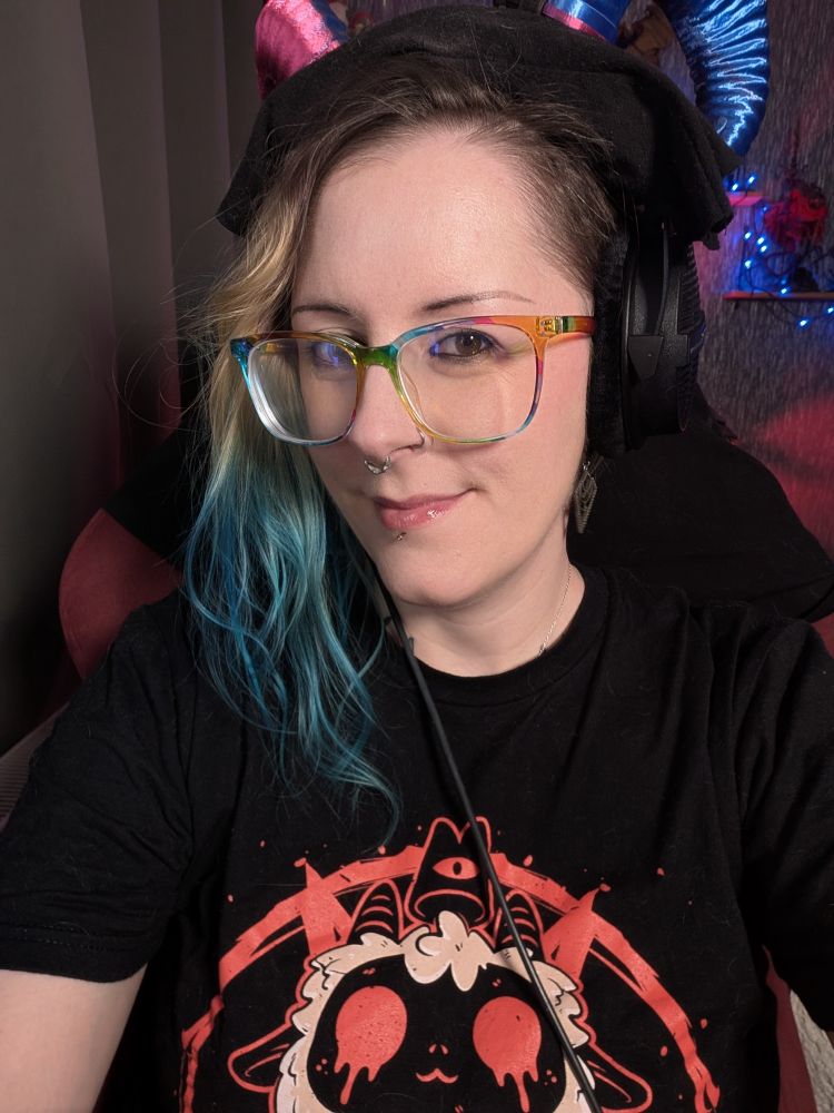 Selfie of me. Blonde and blue hair in a side part. Wearing rainbow glasses and a gaming headset with pink and blue ram horns. Got a cult of the lamb t-shirt on. Smiling at the camera.