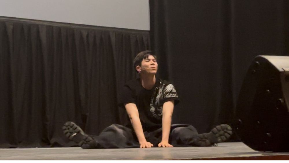 Chanyeon from Big Ocean in all black sitting on a stage with his legs out and his hands in the middle pressed against the stage while he looks slightly up