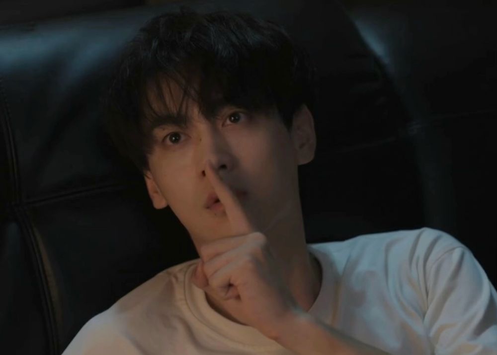 Tain in a white tee leaning on a black headboard while shushing the camera