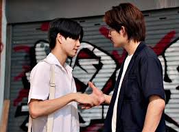 Two men shaking hands in front of a graffitied garage door. The man in the left is in a white button up and white bag, the man on the right is in a white tee with a dark blue short sleeve button up un buttoned over top.