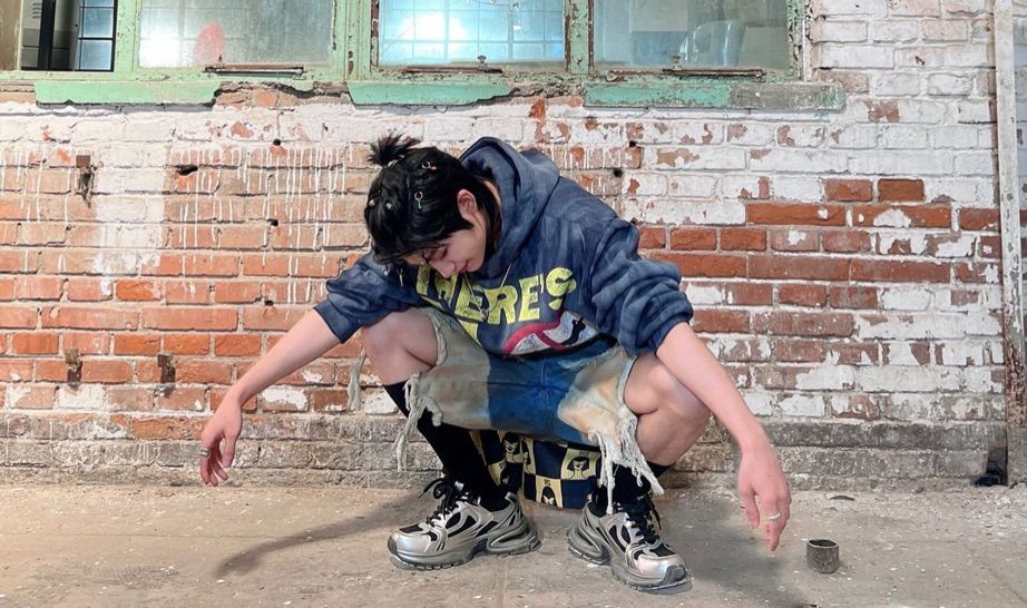 林展烁 from ENONE squatting down while in what and blue jeans with holes at the knee exposing most of his legs. He has silver shoes and black socks on. He has a blue hoodie with yellow words and a white red and black design on it. He has his black hair pulled back and charms attached to it. He is standing on plain concrete in front of half white painted brick wall with warn down green windowsills 