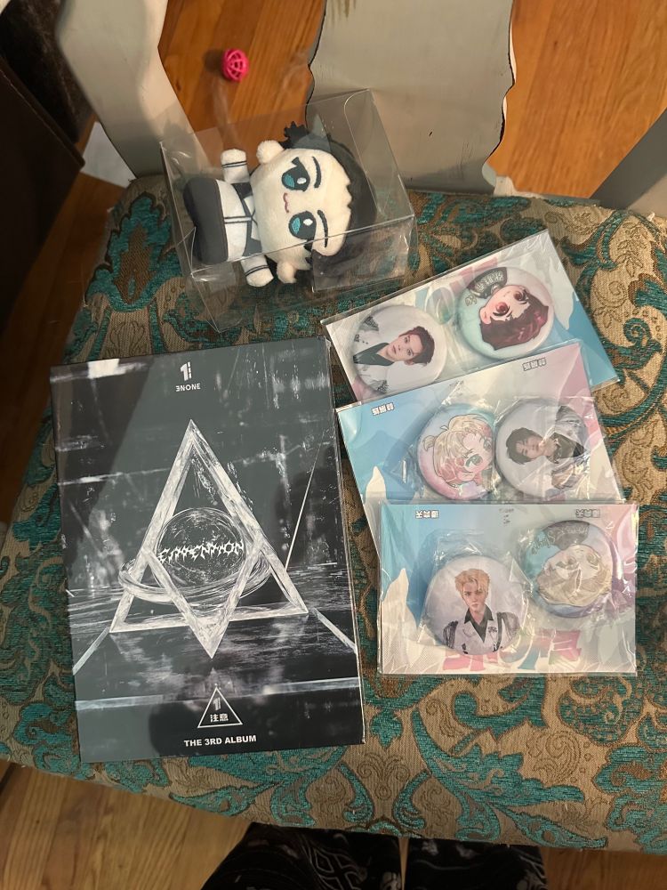 CPop Enone Merch laid out on a green and gold fabric chair on wooden floors. The top clear box has a plush of 林展烁 as a chibi in a white top and black pants. To the right is a black album with white detailing and a White triangle in the middle reading Ettintion and the Enone logo. To the right is three sets of two pins with each member one pin is a photo and the other is a chibi drawing. 