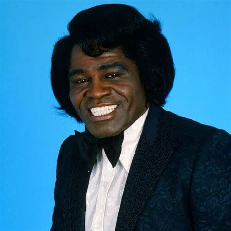 James Brown centered on a sky blue background, wearing a midnight blue with black detailing dinner jacket, white shirt and black bow tie smiling at the camera.