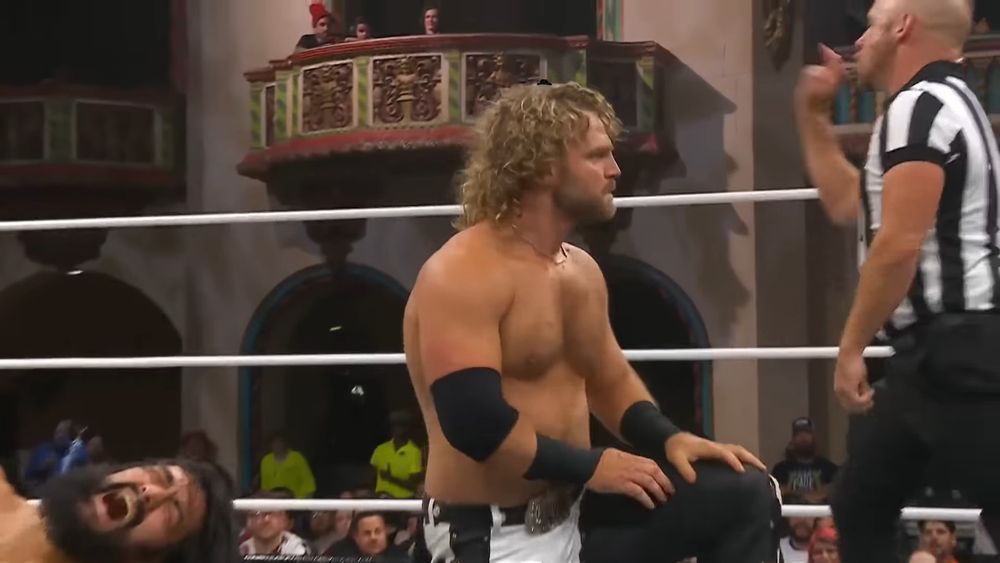 Hangman Adam Page on one knee in the ring during a match, he has a tiny black cartoon cowboy hat photoshopped onto his head. Yuta is dying in the corner but this ain't about him.
