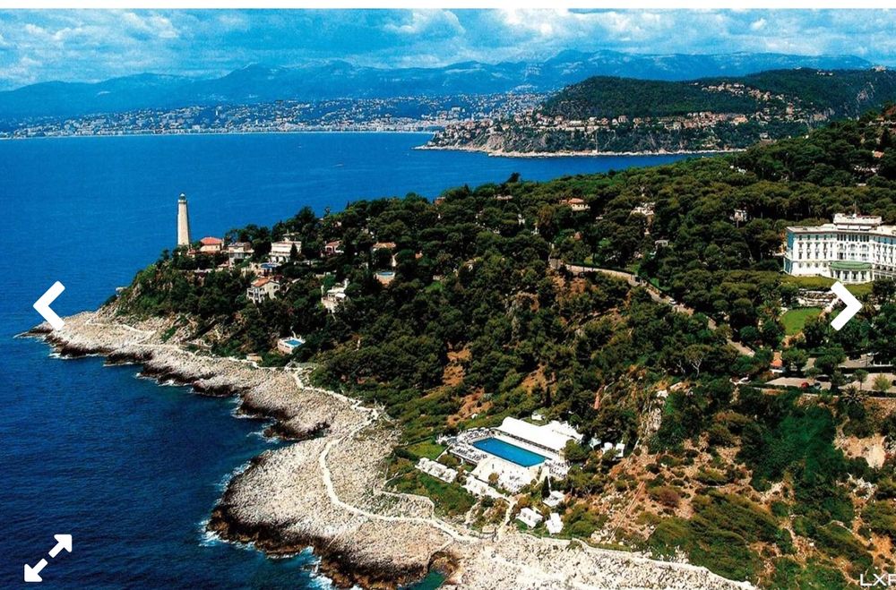 Image of Grand-Hôtel du Cap-Ferrat atop a hill in the French Riviera. The image shows the coast with a lighthouse in the distance and a large pool at the bottom of the hill. 