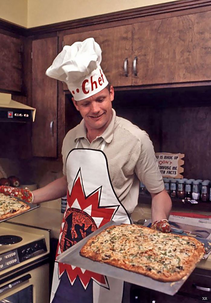 Color photograph of Neil Armstrong wearing oven mitts, an apron and tall chef's hat, holding two squarish pizzas on oven pans. Inside a smallish 1960s domestic kitchen with dark wooden cabinetry and a well supplied spice rack. Oven, counters, and walls are sort of a light olive drab. Chef's hat says "..Chef", apron "1st [occluded]". 
There is a a sign on the wall by the spices that appears to be jokey, but is mostly occluded:
"-- is made up
     DON'T
--onfuse me
--th facts"