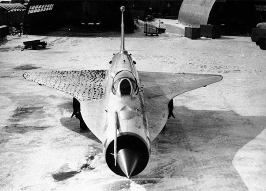 overhead front view of Mig-21 modified with compound delta wing, one wing is covered in small tell tales(fabric strips attached to surface) to study airflow over the wings in flight. black and white, grainy, appears to be outside half-cylindrical hangers in snow