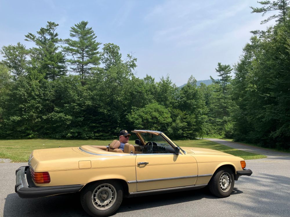 A vintage yellow-gold Mercedes Benz 450SL with beige interior, top down, getting ready to pull out a long driveway. A large lawn and trees along the road are in back of the frame. 