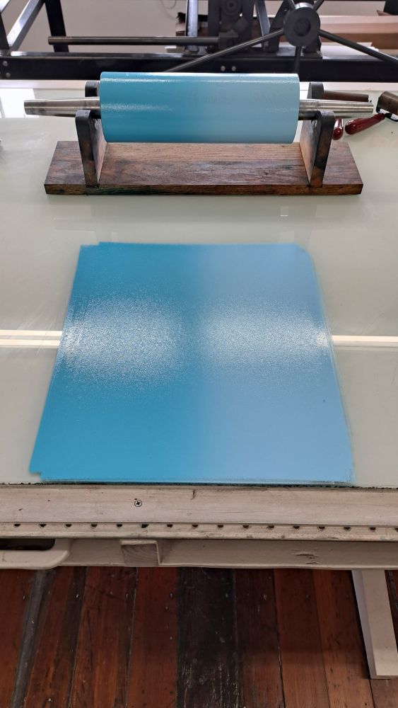 A printer's ink roller sitting in a cradle on a table with glass. On the glass is rolled out ink, in a gradient going from a light to a medium blue. 