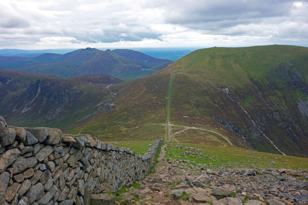A landscape photograph. The left foreground of the shot is dominated by a drystone wall that snakes down the slope of a very steep hill and up another. A saddle between two hills is in the exact centre of the shot, with a steep, rocky gorge visible to the left of the shot and another hill to the right. The landscape is green, but bleak. Not a single person is visible in the shot. In the distance, to the left of the shot, a mountain with two rocky colls on the summit can be seen. Dark clouds loom in the sky. 