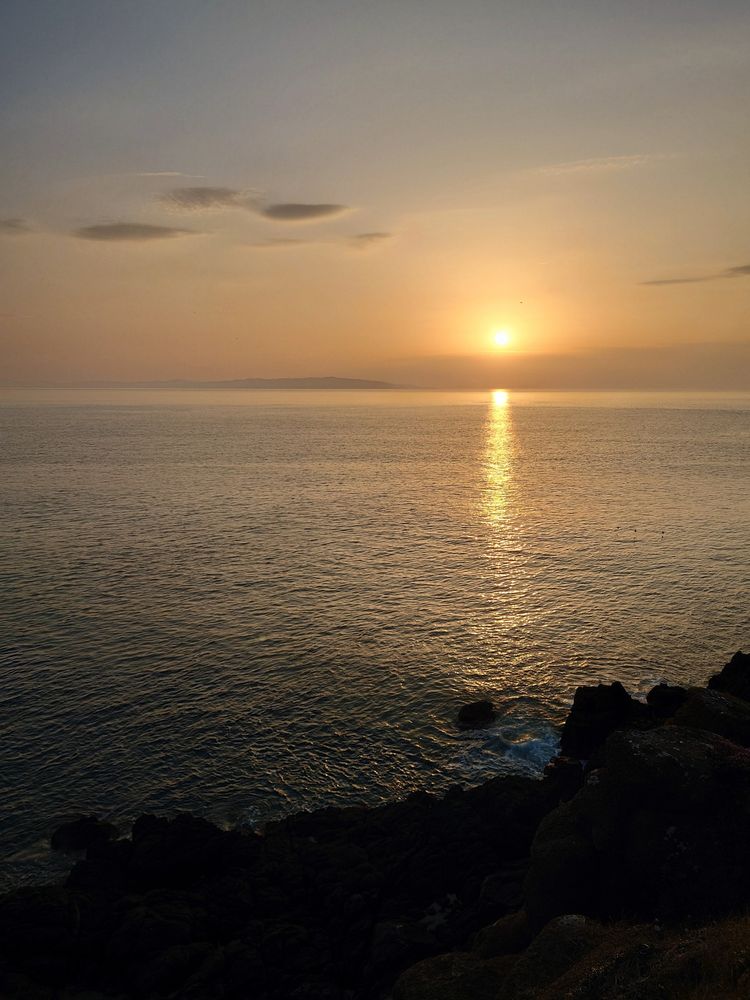A photograph of a sunset. The sun is just above the horizon, casting a long ray of shimmering light across the ocean towards the foreground. The light is muted but has a quality of soft radiance. The foreground is a rocky shore, and a distant coastline sits on the horizon. A few clouds are visible in an otherwise clear sky, the sky's colour transiting from light blue to a soft red above the horizon.
