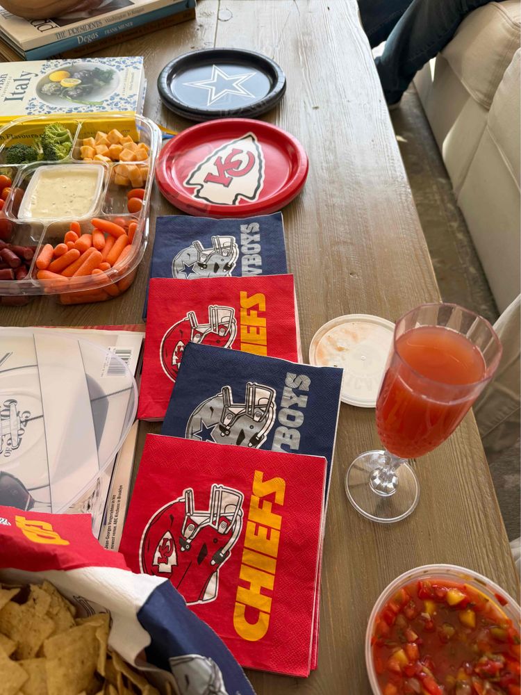 Stacks of Chiefs and Cowboys napkins and plates sitting next to each other amongst trays of snacks. 