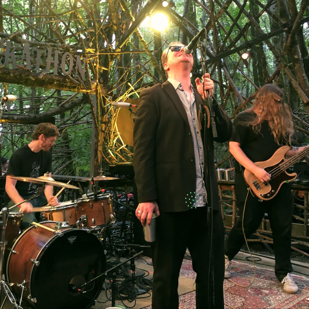 Up close photo of three members of Protomartyr (left to right, Alex Leonard on drums, Joe Casey singing, and Scott Davidson on bass). THey are playing on a stage with a wooden thicket in back of them and the whole stage is in the woods (called the Woods Stage). 