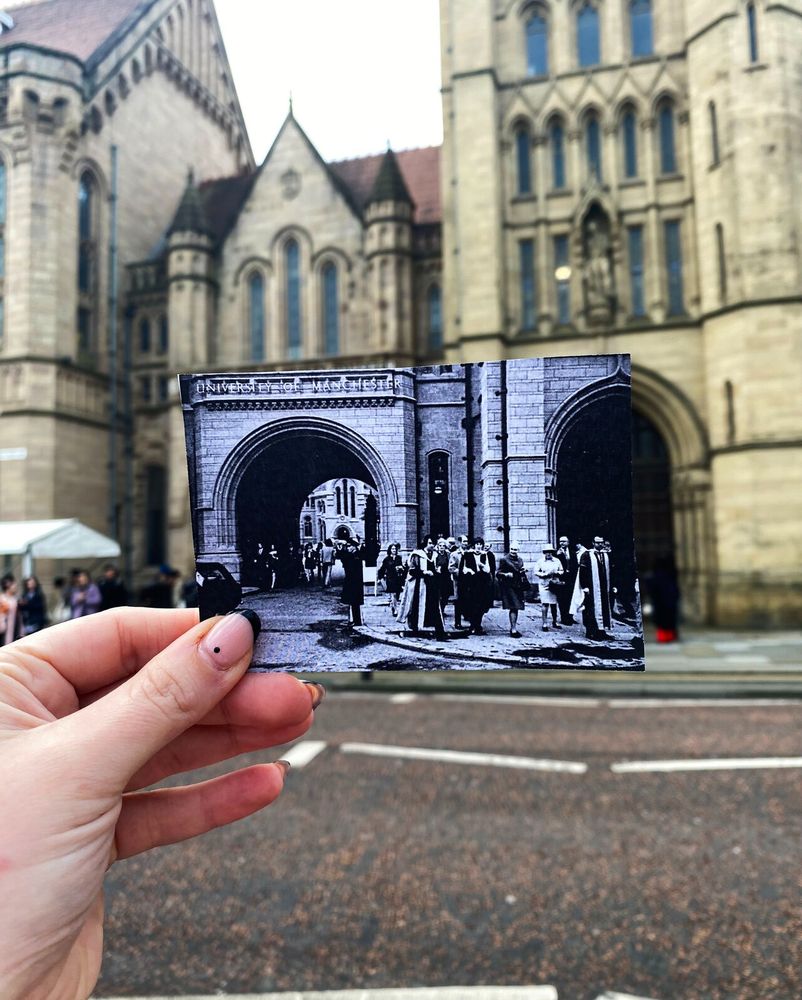 A hand is holding up a black and white photo of an old graduation ceremony outside the Queen's Arch on Oxford Road, in front of the arch so that it lines up to present day