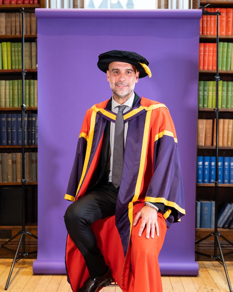 Pep Guardiola, manager of Manchester City Football Club, sits in front of a purple backdrop with book cases either side. He wears a dark purple, red and gold robe and black cap after being awarded an honorary degree by The University of Manchester.